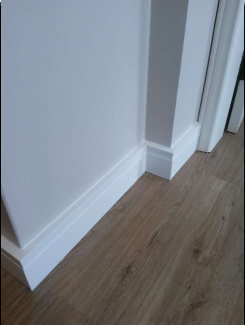 Detalhe do encontro entre o rodapé branco e o piso vinílico em carvalho, com o canto externo fechado