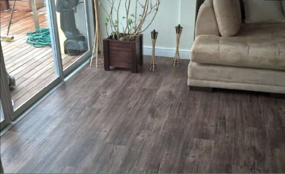 Sala de estar com piso vinílico em tom carvalho fumê seguindo até a porta de vidro da varanda